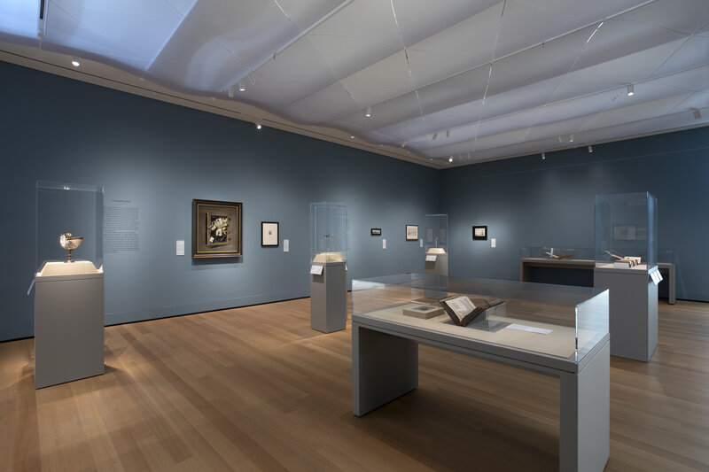View of a blue-walled exhibition with objects in cases in the foreground and paintings on the walls in the distance