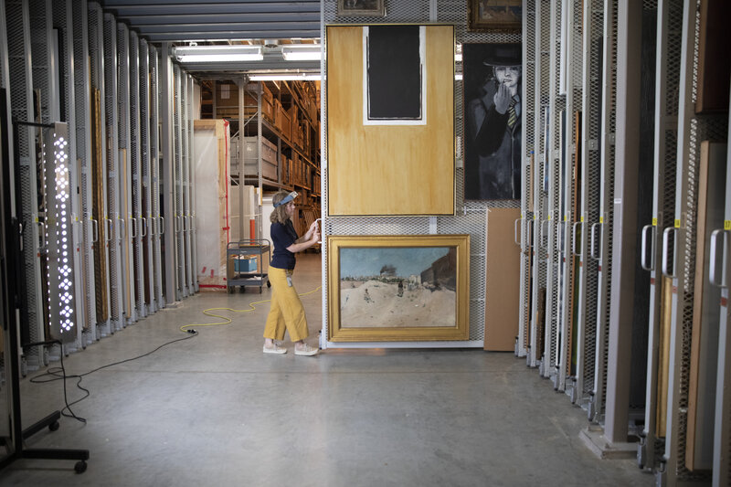 Women in dark top and yellow pants moves a storage wall that holds several paintings.