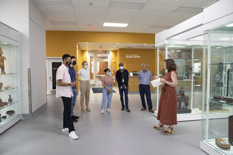 A group of people stand in a semi-circle in discussion in a room with glass cases filled with art objects