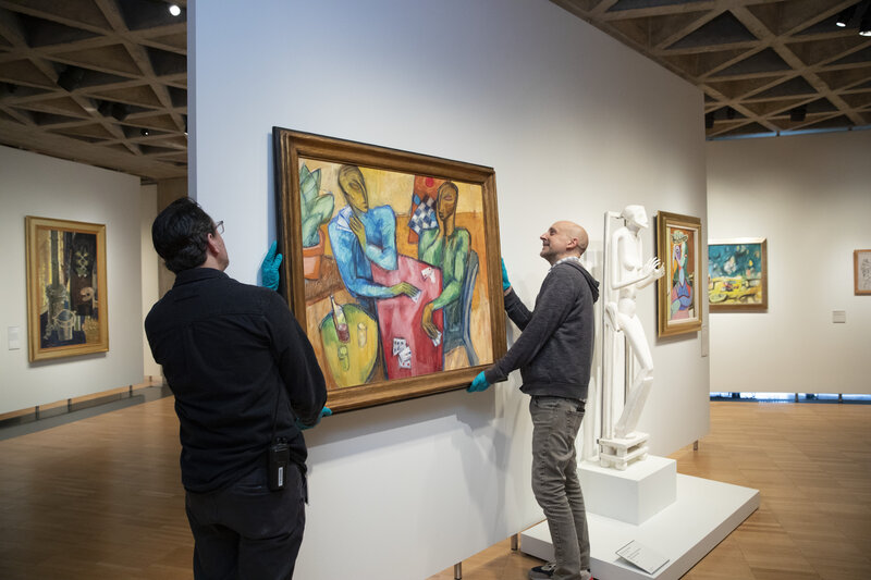 Two men wearing green gloves stand on either side of a painting, holding it as though they are removing it from the wall. Other art objects are on the walls in the room