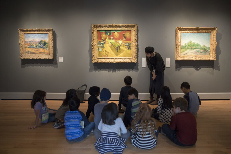 A group of children sit on the floor of a gallery looking at three paintings on a grey wall. A teacher stands by the paintings, bending down to the children in conversation