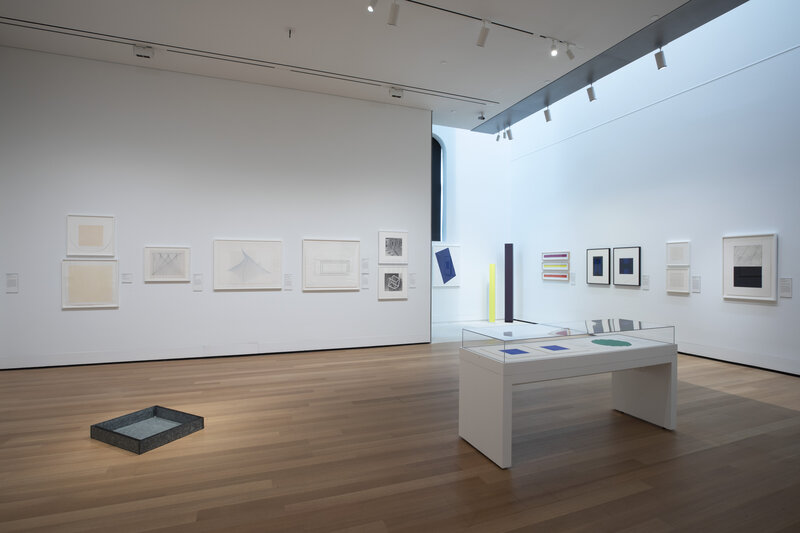 A view of an installation in a gallery with wooden floors and white walls and ceilings. Artworks hang on the walls, and a vitrine containing additional works stands in the right middle ground. Two works of sculpture are visible, one in the left middle ground and the other in the center background. Both are geometric in shape.