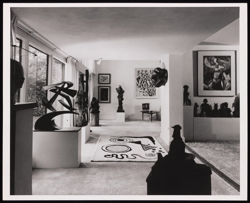 A black-and-white photograph of the carpeted interior of a home. Artworks appear throughout the space. Some hang on the wall, while others are works of sculpture presented on platforms or in vitrines. Light passing through large glass doors or windows at left illuminates the space.  