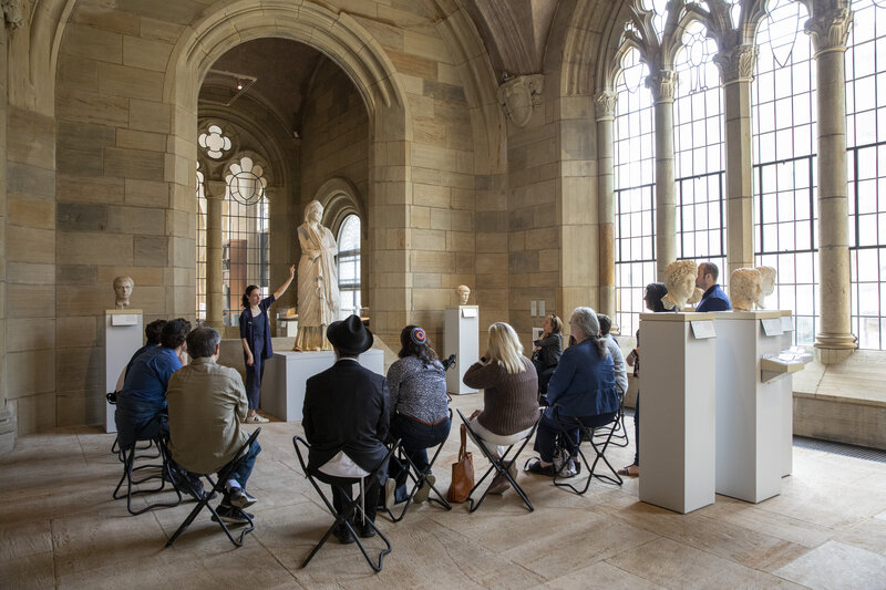 A group of people seen from behind as they sit on folding chairs in a bright gallery space. They look toward a standing person, who gestures at a large figural sculpture presented on a pedestal.