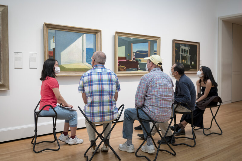 Six people seen largely from behind as they sit on folding chairs in a gallery space. The people wear surgical masks. They look toward a painting that hangs on a white wall alongside other artworks of similar scale. 