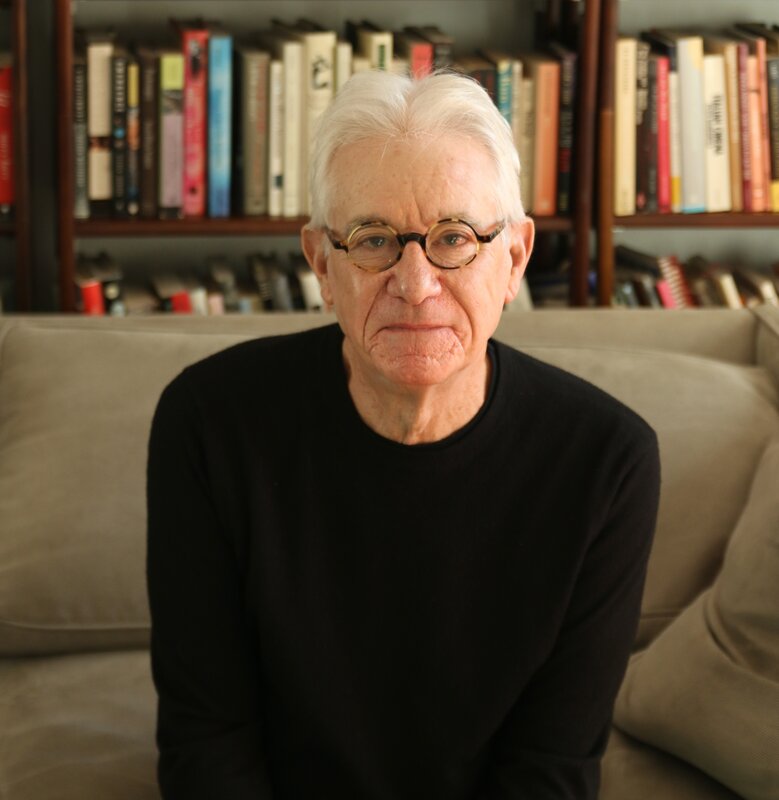 A seated man seen from the waist up. He looks into the camera with a gentle expression. He has short white hair and wears glasses with round, tortoise-shell frames, along with a black, long-sleeve top. 