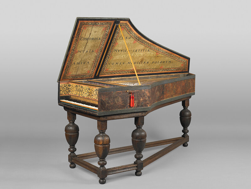 A keyboard instrument resembling a piano. Text and ornamentation are visible on the inside of its raised lid and on the surfaces above the keyboard itself. The instrument has elaborately carved legs.