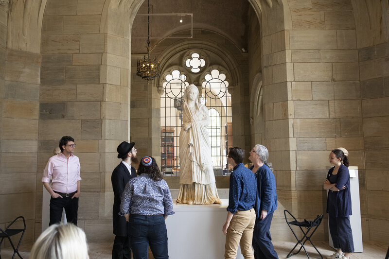 A group of people stands gathered around a large figural sculpture presented on a pedestal. In the background, a tall window with tracery elements looks into another space. 