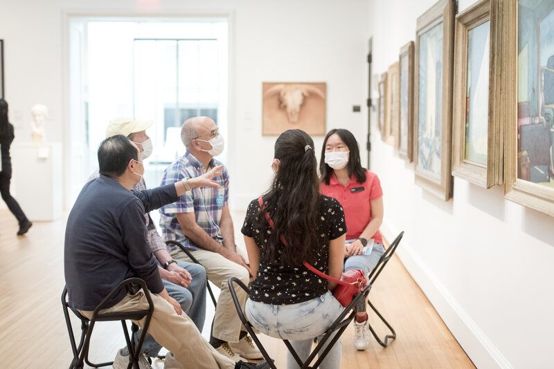 Five people sit on folding chairs in a gallery space. The people wear surgical masks. They look toward a painting that hangs on a white wall alongside other artworks of similar scale. 