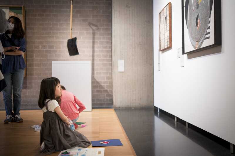 A child seen in profile while kneeling on the floor of a gallery. She looks toward a work of art that hangs on the wall in front of her. Behind her, a shovel, perhaps a work of sculpture, hangs from the ceiling.  