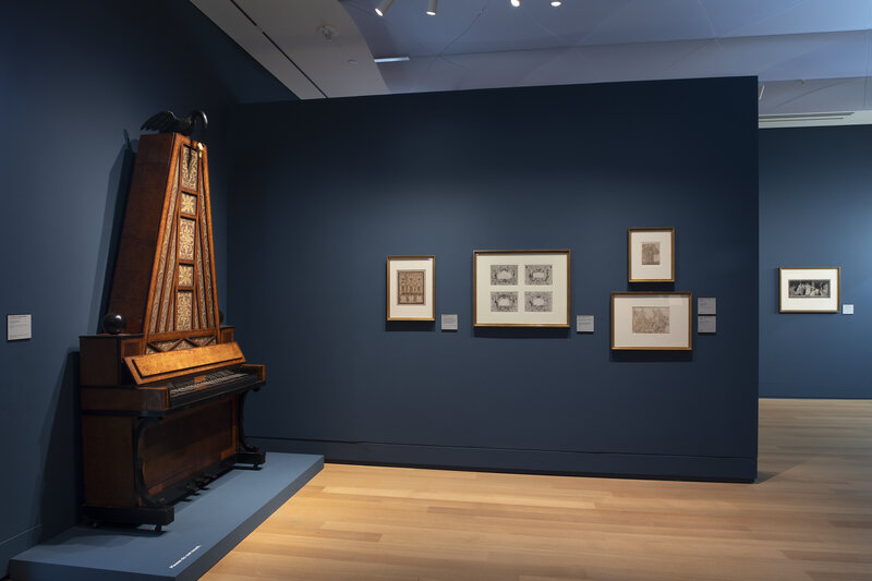 A gallery installation with midnight blue walls, on which hang framed works of art. On a platform at left is an object resembling a piano. A triangular segment rising vertically from its keyboard features ornamentation.