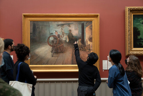 A group of young women and men examine Henry Ossawa Tanner's painting Spinning By Firelight.