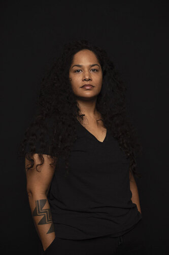 A woman seen from the waist up. She faces the viewer and has her hands in her pockets. She is dressed all in black, with a V-neck T-shirt. Her long, curly black hair falls to her chest. A tattoo consisting of geometric shapes is visible on her right forea