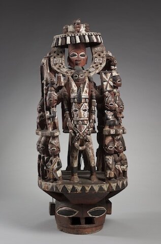 Carved wooden mask depicting a richly adorned ruler on horseback carrying a sword and wearing protective amulets on his arms. Three tiers of small-scale attendant figures surround the ruler.